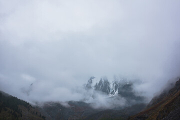 Dark atmospheric landscape with high snow mountains in dense fog in rainy weather. Large snowy mountain range in thick fog in dramatic overcast. Snow-covered rocky mountains in low clouds during rain.