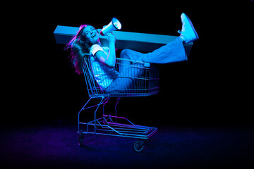 Cheerful astonished girl sitting in shopping cart and inviting for shopping, black friday, seasonal sales isolated over dark background in neon light. © master1305