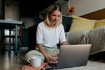 Fototapeta premium Blond woman wearing white t-shirt and light pants sitting on the floor at home and working on laptop with coffee. Adorable girl working remote at home