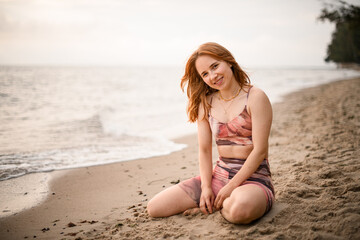 Great view on caucasian smiling beautiful red-haired young woman sitting on sand of beach.