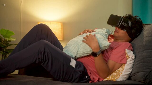 Tired young man with VR or virtual reality goggles sleeping on couch at home during night - conept of addiction, relaxation and futuristic.