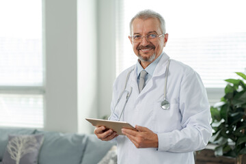 Portrait of an elderly doctor, he looks into the camera. The doctor stands against the background of a medical clinic and holds a tablet in his hands.