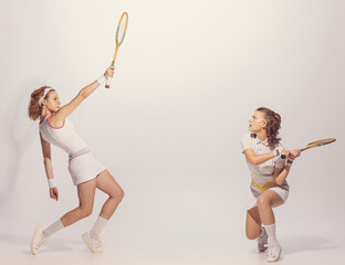 Portrait of two beautiful young women in retro uniform playing badminton isolated over grey studio background