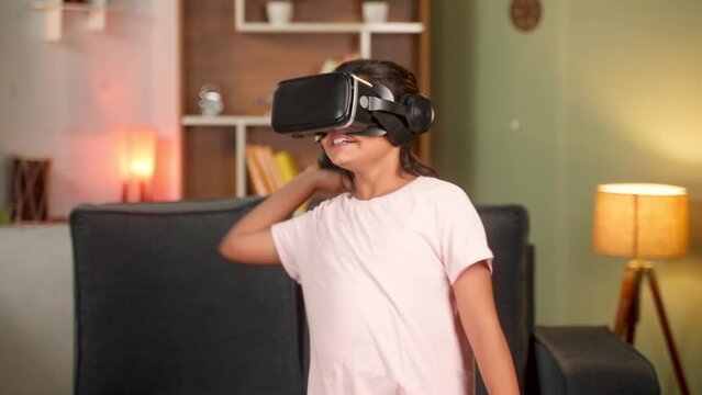 handheld shot of Girl child with VR or vertual reality goggles playing video game on metaverse using joystick at home - concept of immersive experience, technology and entertainment.