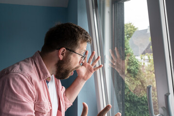 Surprised man looks out the open window, he is shocked by what he saw, opens mouth and emotionally makes a gesture with hands. 