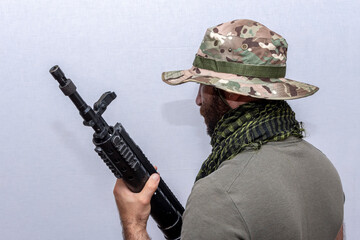 A man in an army panama hat and a military T-shirt holds a rifle, turned sideways to the camera, light background. Concept: soldier of fortune, mercenary service in the army.