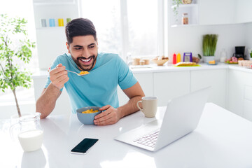 Photo of positive handsome guy hold spoon eat healthy cornflakes watch comedy video netbook free time morning indoors