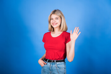 Fototapeta na wymiar Young and attractive caucasian blonde girl in casual clothes waving her hand and greeting isolated on blue studio background.
