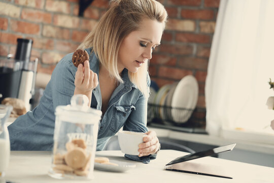 Image of an attractive woman holding digital tablet and eating a cookie in the kitchen at home