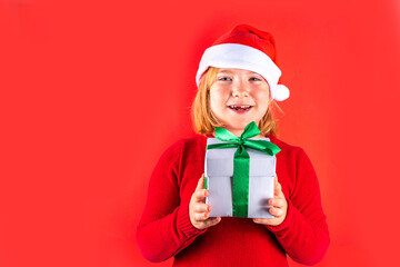 Christmas holiday background with funny blond girl. Little kid girl in red Christmas sweater and funny santa hat costume, with gift box on red background, Xmas greeting card, copy space