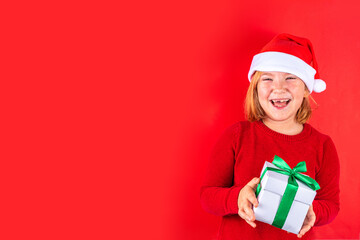 Christmas holiday background with funny blond girl. Little kid girl in red Christmas sweater and funny santa hat costume, with gift box on red background, Xmas greeting card, copy space