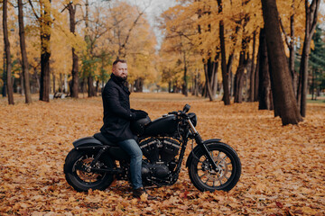 Outdoor shot of male biker with thick beard, wears protective gloves, black coat and jeans, poses on motobike in beautiful park with orange leaves, holds helmet, stops to have rest. Urban lifestyle