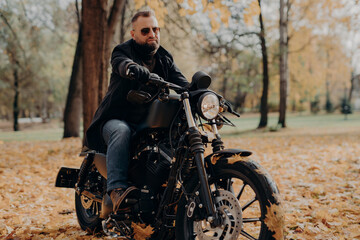 Fototapeta na wymiar Male motorcyclist drives in nature on fast bike, wears shades, black jacket, gloves, jeans and boots, enjoys autumn season, spends free time actively, ready for long trip. People, transport, driving