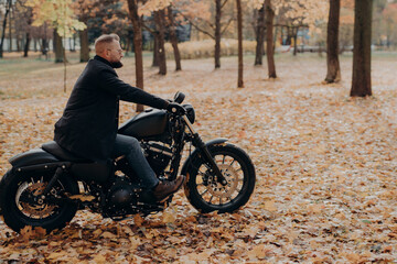 Fototapeta na wymiar Horizontal shot of bearded biker in action or movement against park during autumn time, going to reach destination quickly, wears sunglasses, jacket, jeans and shoes, has adventurous trip alone