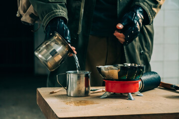 Male stalker in protective suit pours boiled water into cup. Preparation of drinking water using gas cylinder and portable burner. Survival after disaster or war.