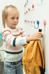 Cute little caucasian girl hanging yellow backpack on a white hanger in the school locker room. Back to school concept