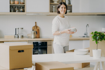 Happy brunette woman in casual outfit, moves into new house, unpackes boxes with kitchen dishes, holds plates, stands near table, green indoor plan in pot, enjoys domestic atmosphere. Modern apartment