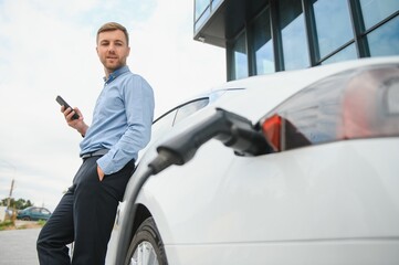Man charging his electric car at charge station and using smartphone