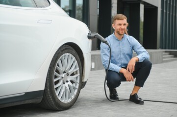 Fototapeta na wymiar Hansome bearded guy sitting near his new modern electric car and holding plug of the charger, while car is charging at the charging station.