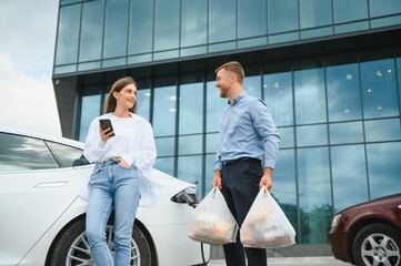 Happy young adult man and smiling woman charging electric car, making shopping and packing bags with food in automobile trunk. Two friends talking and spending free time together
