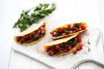 Three traditional fresh Mexican tacos with sliced tomatoes and greens on a white cutting board