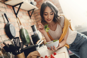 A young, beautiful, charming woman eats fresh vegetable salad in the kitchen at home