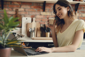Young, charming, brunette girl working on the computer and holding a cup of coffee in the kitchen...