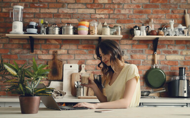 Young, charming, brunette girl working on the computer and holding a cup of coffee in the kitchen at home