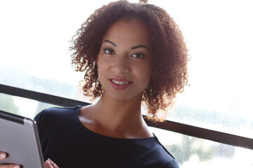 Close-up image of a beautiful woman with curly hair holding digital tablet in front of window