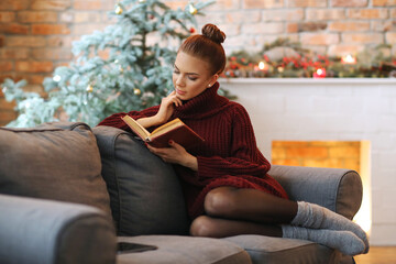 Photo of a cute, pretty girl with a bun on her head in a cozy sweater sitting on the couch with an...