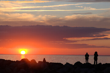 Sunset at Cape Cod beach. Dark outlines (silhouettes) of unrecognizable people on breakwater rocks viewing colorful sky and yellow sun just above horizon with reflection on water.