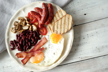 Top view of English breakfast with fried eggs, bacon,sausages, bread and vegetables in white plate.