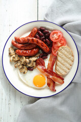 Close view of English breakfast with fried eggs, bacon,sausages, bread and vegetables in white plate.