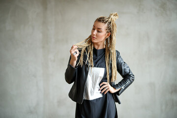 Photo of a young woman with dreadlocks wearing leather jacket standing and posing against stone background.