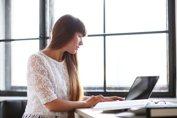 Beautiful, sweet, young girl with long hair sitting and working on the computer against windowsill.