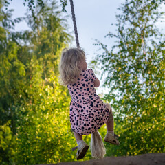 A baby girl in a pink dress swings on a bungee swing made of a wooden board with a rope attached to...