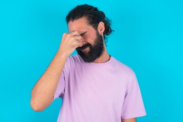 Sad young bearded man wearing violet T-shirt over blue studio background suffering from headache holding hand on her face