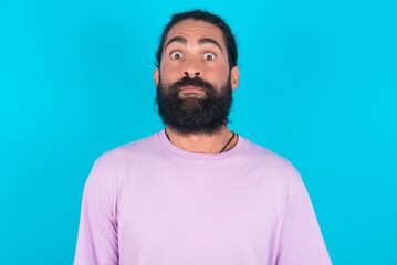 Stunned young bearded man wearing violet T-shirt over blue studio background stares reacts on shocking news. Astonished latin man holds breath