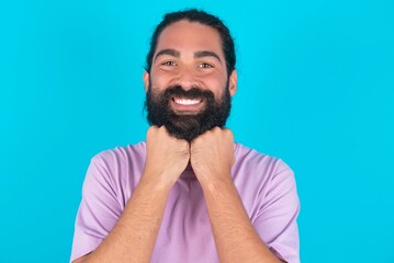 Satisfied young bearded man wearing violet T-shirt over blue studio background touches chin with both hands, smiles pleasantly, rejoices good day with lover