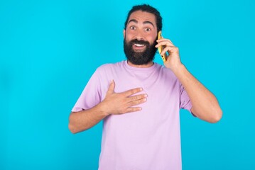 Smiling young bearded man wearing violet T-shirt over blue studio background  talks via cellphone, enjoys pleasant great conversation. People, technology, communication concept