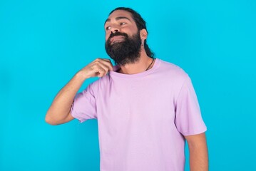 young bearded man wearing violet T-shirt over blue studio background stressed, anxious, tired and frustrated, pulling shirt neck, looking frustrated with problem