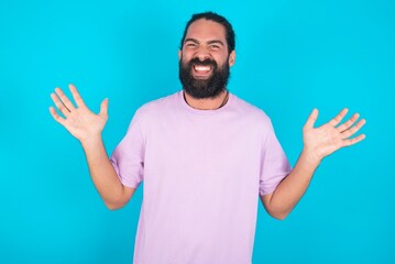 Crazy outraged young bearded man wearing violet T-shirt over blue studio background screams loudly and gestures angrily yells furiously. Negative human emotions feelings concept