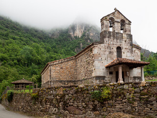 Iglesia de Pola de Somiedo de piedra con campanario, sobre un fondo verde con niebla en las montañas, en verano de 2021, España.