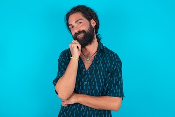 Cheerful young bearded man wearing blue shirt over blue studio background with hand near face. Looking with glad expression at the camera after listening to good news. Confidence.