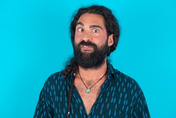 young bearded man wearing blue shirt over blue studio background crosses eyes, puts lips, makes grimace with awkward expression has fun alone, plays fool.