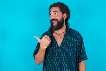 Stupefied young bearded man wearing blue shirt over blue studio backgro with surprised expression, opens eyes and mouth widely, points aside with thumb, shows something strange. Advertisement concept.
