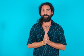 young bearded man wearing blue shirt over blue studio background keeps palms pressed together in...