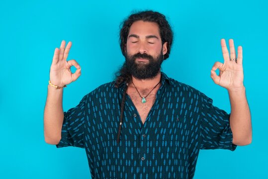 young bearded man wearing blue shirt over blue studio background doing yoga, keeping eyes closed, holding fingers in mudra gesture. Meditation, religion and spiritual practices.