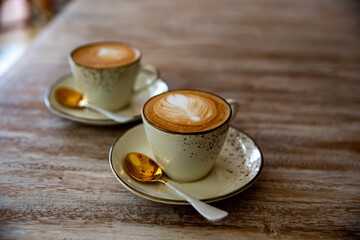 Two stylish white cups of cappuccino with latte art on saucer and golden spoons on light wooden table.