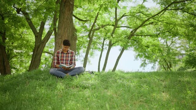 African American teenager reading a book under tree in city park, education